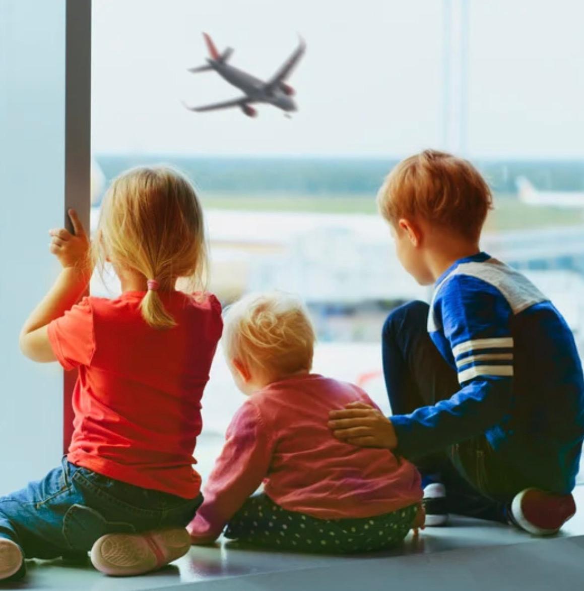 Trois enfants assis devant la baie vitrée de l'aéroport regardent un avion décoller